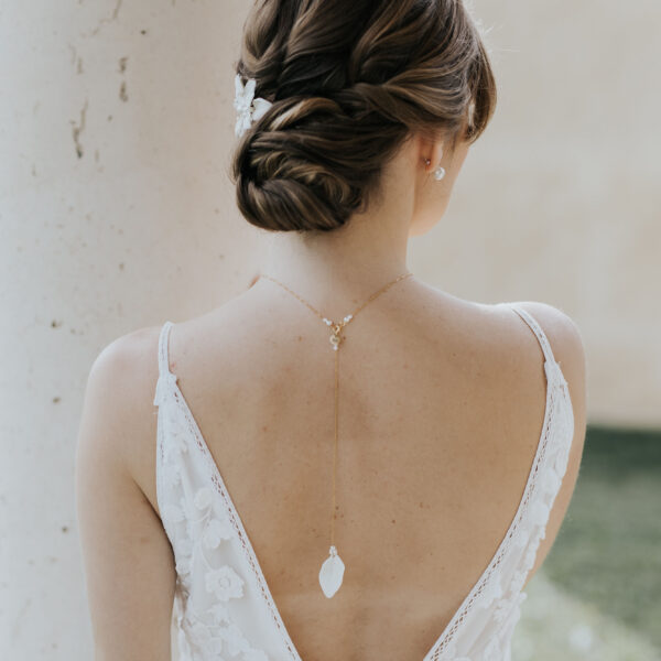 Jeune mariée portant une robe dos nu et des bijoux de mariage en porcelaine. Elle porte un pendentif de dos et dans ses cheveux, un ornement floral en porcelaine.