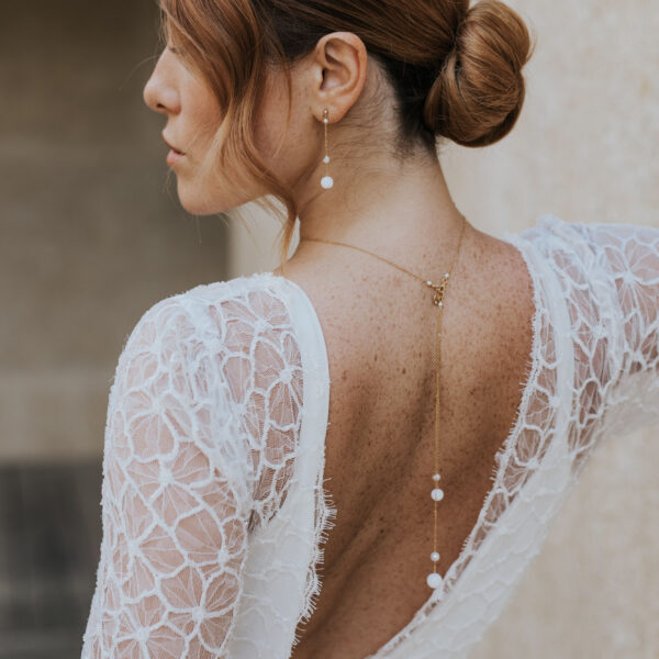 Bijoux de mariage élégants et minimalistes en perle d'eau douce et porcelaine, portés par un mariée. Elle porte des boucles d'oreilles pendantes et sur une robe dos nu, un pendentif de dos.