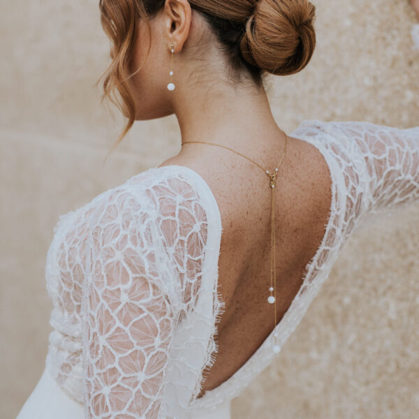 Bijoux de mariage élégants et minimalistes en perle d'eau douce et porcelaine, portés par un mariée. Elle porte des boucles d'oreilles pendantes et sur une robe dos nu, un pendentif de dos.