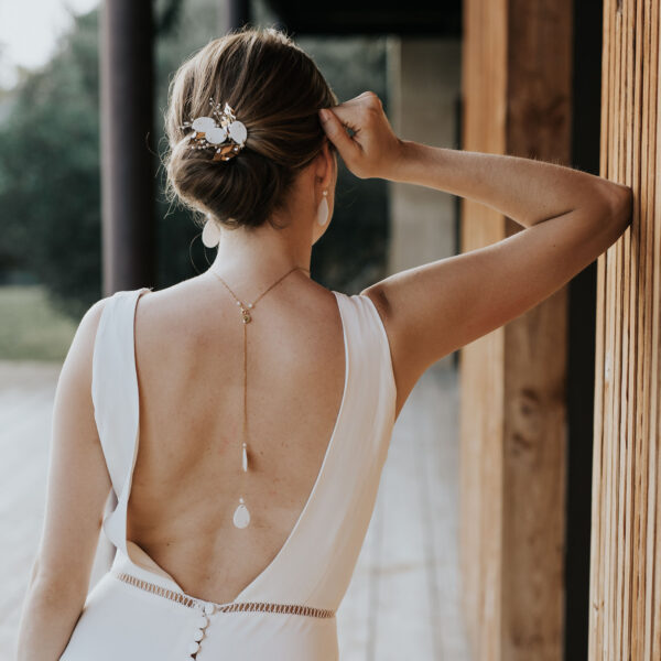 Mariée portant ses bijoux de mariage en porcelaine minimalistes et élégants. Elle porte des boucles d'oreilles et un pendentif de dos sur sa robe dos nu et des épingles à chignon dans sa coiffure de mariée.