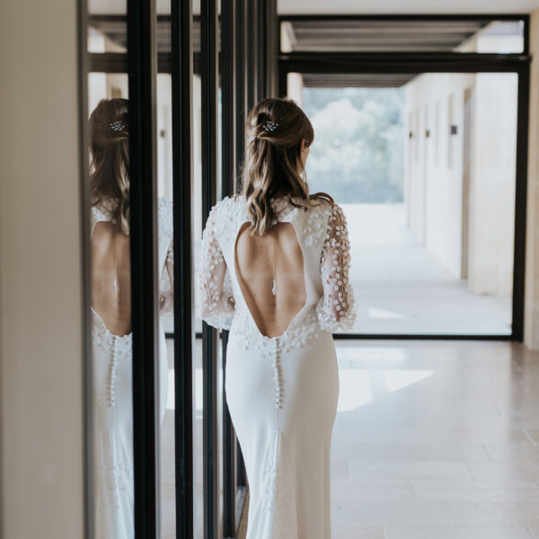 Une mariée de dos, adossée à une vitre, porte une robe dos nu habillée d'un pendentif de dos. Ses cheveux sont lachés et elle porte un bijou de mariage dans sa coiffure.